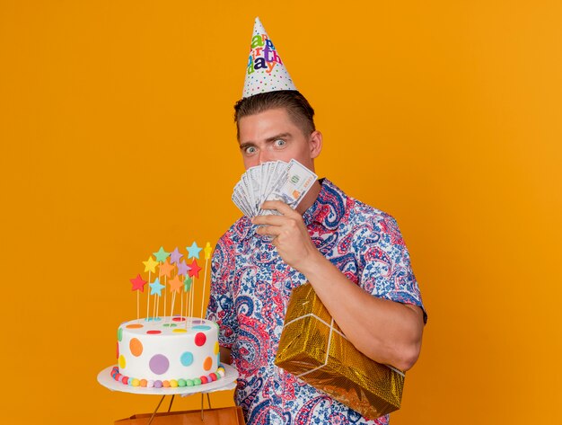 Impressionné jeune homme de fête portant un chapeau d'anniversaire tenant des cadeaux avec un gâteau et le visage couvert avec de l'argent isolé sur orange