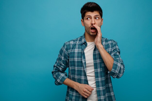 Impressionné jeune homme gardant la main près de la bouche regardant côté chuchotant isolé sur fond bleu avec espace de copie