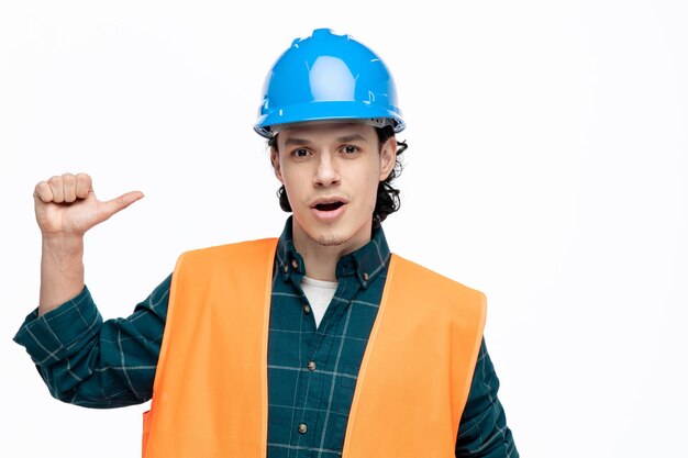 Impressionné jeune homme ingénieur portant un casque de sécurité et un gilet de sécurité regardant la caméra pointant vers lui-même isolé sur fond blanc