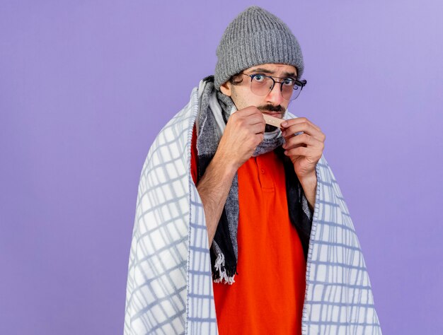 Impressionné jeune homme malade de race blanche portant des lunettes chapeau d'hiver et écharpe enveloppé dans un plaid tenant du plâtre médical devant le menton isolé sur un mur violet avec espace de copie