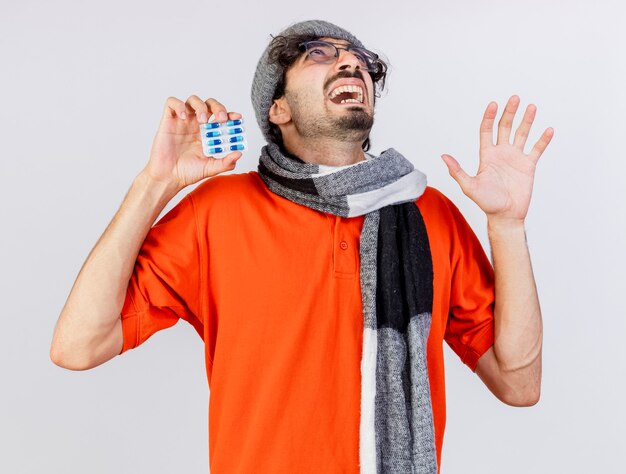 Impressionné jeune homme malade de race blanche portant des lunettes chapeau d'hiver et écharpe montrant pack de capsules et main vide en levant isolé sur fond blanc