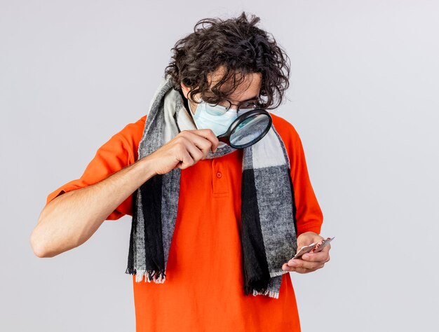 Impressionné jeune homme malade de race blanche portant des lunettes foulard et masque tenant des pilules médicales en les regardant à travers une loupe isolé sur fond blanc avec espace copie