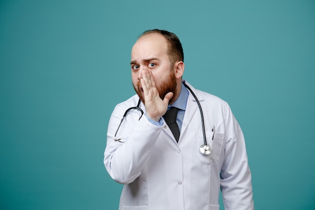 impressionné jeune homme médecin portant un manteau médical et un stéthoscope autour du cou en gardant la main près de la bouche en regardant la caméra en chuchotant isolé sur fond bleu