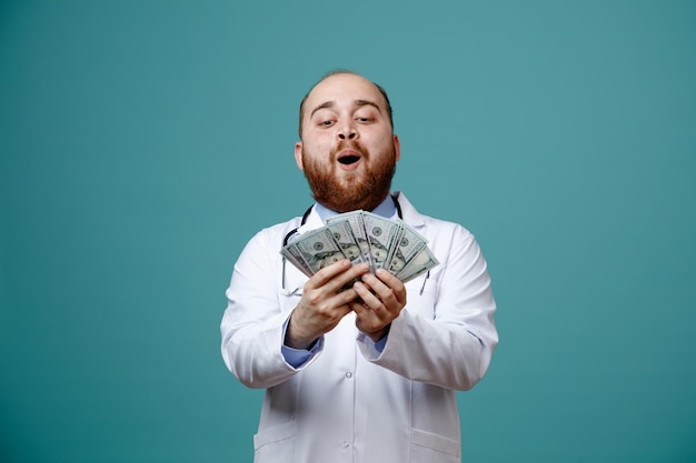 Impressionné jeune homme médecin portant un manteau médical et un stéthoscope autour de son cou tenant et regardant de l'argent isolé sur fond bleu