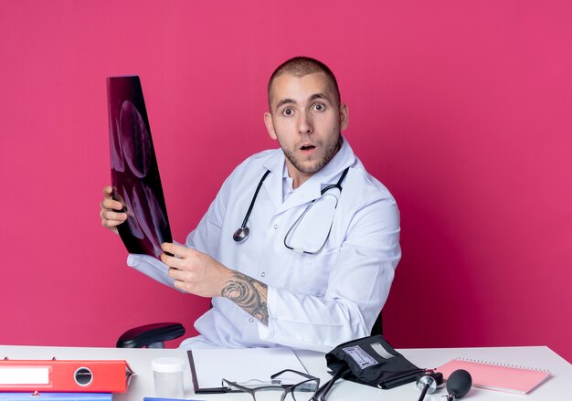 Impressionné jeune homme médecin portant une robe médicale et un stéthoscope assis au bureau avec des outils de travail tenant des rayons x à l'avant isolé sur rose