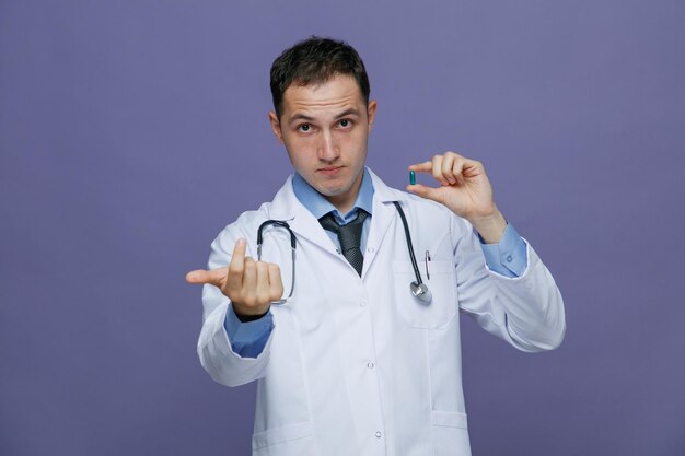 Impressionné jeune homme médecin portant une robe médicale et un stéthoscope autour du cou regardant la caméra montrant la capsule faisant venir ici le geste isolé sur fond violet