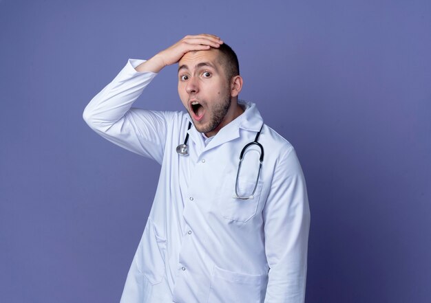 Impressionné jeune homme médecin portant une robe médicale et un stéthoscope autour de son cou mettant la main sur la tête isolé sur violet