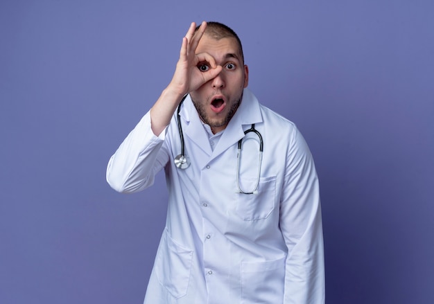 Impressionné jeune homme médecin portant une robe médicale et un stéthoscope faisant regard geste isolé sur violet