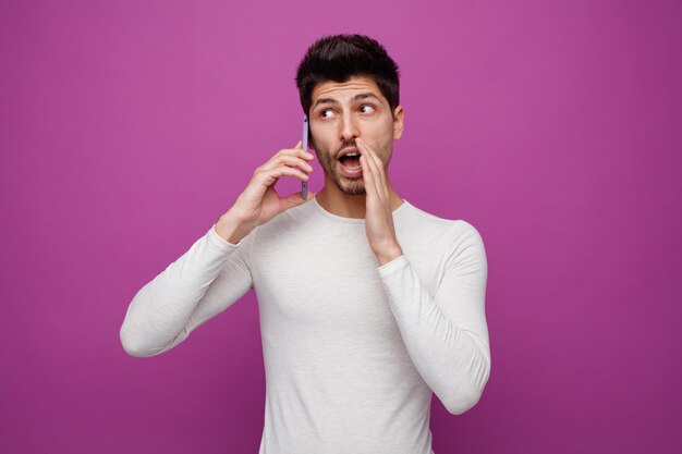 Impressionné jeune homme parlant au téléphone en gardant la main près de la bouche en regardant le côté chuchotant isolé sur fond violet