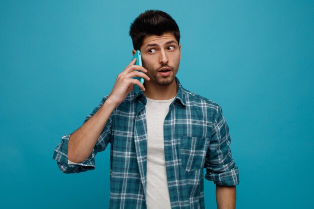 Impressionné jeune homme parlant au téléphone regardant à côté isolé sur fond bleu