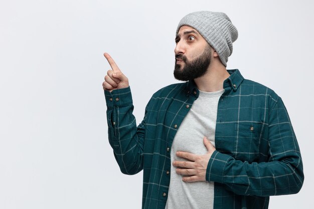 Impressionné jeune homme portant un chapeau d'hiver en gardant la main sur le ventre en regardant le côté pointant vers le haut isolé sur fond blanc avec espace de copie