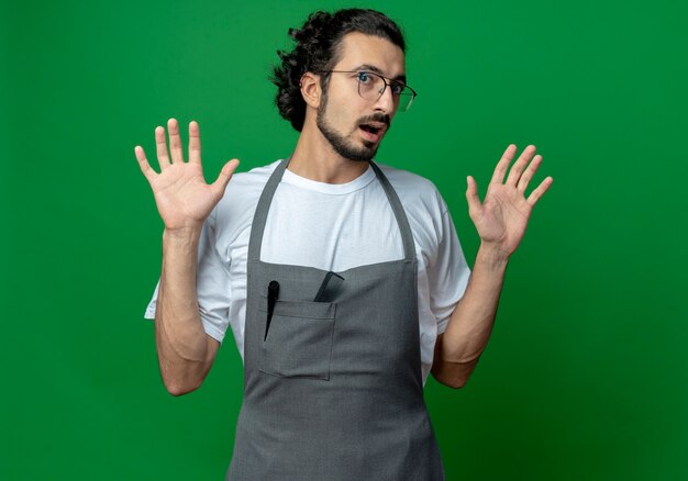 Impressionné jeune homme de race blanche barbier portant des lunettes et bande de cheveux ondulés en uniforme montrant les mains vides