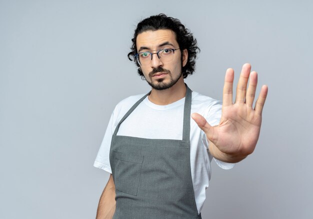 Impressionné jeune homme de race blanche coiffeur portant des lunettes et bande de cheveux ondulés en uniforme gesticulant arrêter à la caméra isolée sur fond blanc avec espace de copie