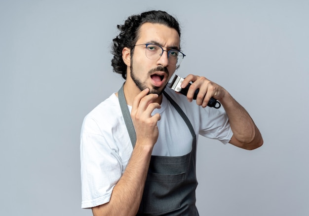 Impressionné jeune homme de race blanche coiffeur portant des lunettes et une bande de cheveux ondulés en uniforme taillant sa barbe avec une tondeuse à cheveux et touchant son menton isolé sur fond blanc avec espace de copie