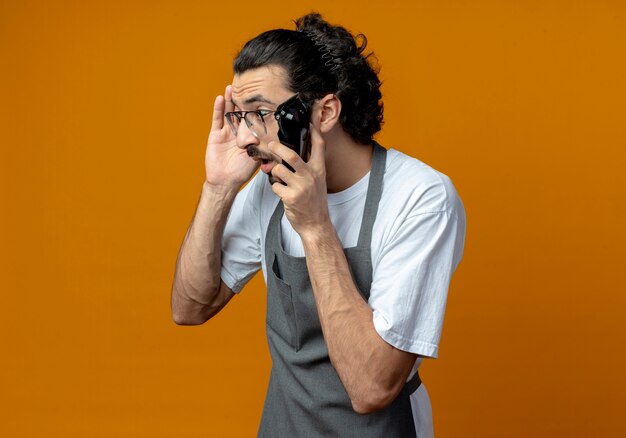 Impressionné jeune homme de race blanche coiffeur portant l'uniforme et des lunettes tenant une tondeuse à cheveux touchant la tête et regardant à côté isolé sur fond orange avec copie espace