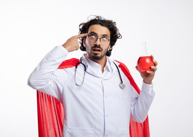 Impressionné jeune homme de race blanche à lunettes optiques portant l'uniforme de médecin avec manteau rouge et avec stéthoscope autour du cou met le doigt sur la tempe et détient un liquide chimique rouge dans une fiole en verre