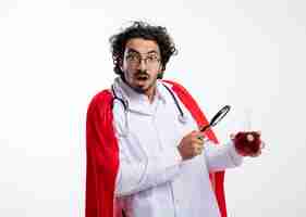 Photo gratuite impressionné jeune homme de race blanche à lunettes optiques portant l'uniforme de médecin avec manteau rouge et avec stéthoscope autour du cou tient une loupe et un liquide chimique rouge dans un flacon en verre