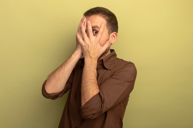 Impressionné jeune homme de race blanche mettant les mains sur le visage en regardant la caméra à travers les doigts isolés sur fond vert olive avec espace copie