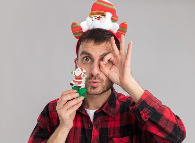 Impressionné jeune homme de race blanche portant le bandeau du père noël tenant le jouet de Noël bonhomme de neige regardant la caméra faisant le geste de regard isolé sur fond blanc