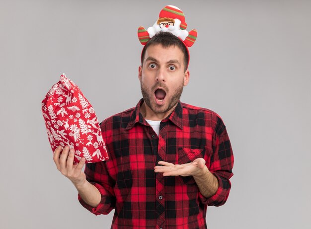 Impressionné jeune homme de race blanche portant bandeau de Noël tenant et pointant avec la main au sac de Noël isolé sur mur blanc avec espace copie