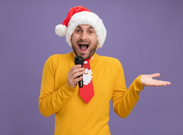 Impressionné jeune homme de race blanche portant chapeau de Noël et cravate tenant microphone montrant la main vide isolé sur mur violet