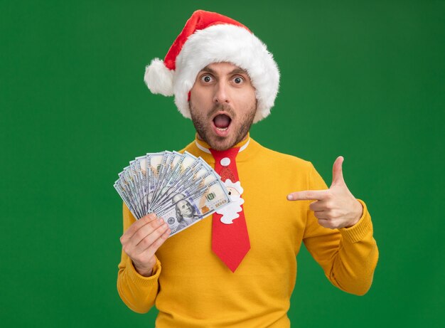 Impressionné jeune homme de race blanche portant chapeau de Noël et cravate tenant et pointant sur l'argent isolé sur mur vert