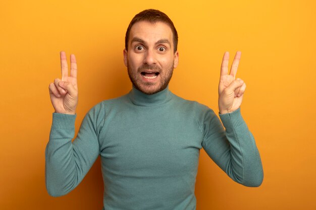 Impressionné jeune homme de race blanche regardant la caméra faisant des signes de paix isolés sur fond orange