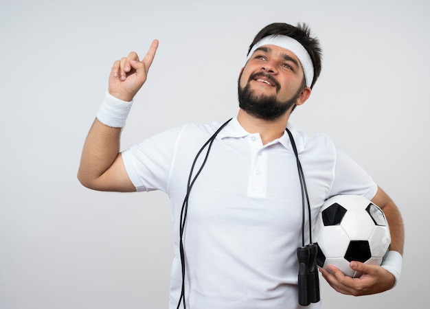 Impressionné jeune homme sportif portant un bandeau et un bracelet avec une corde à sauter sur l'épaule tenant des points à bille en haut isolé sur un mur blanc