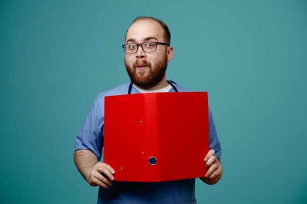 Impressionné jeune infirmier portant des lunettes gommage infirmière et stéthoscope autour de son cou tenant dossier regardant caméra isolé sur fond bleu