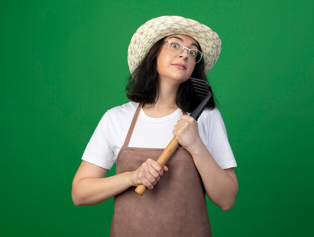 Impressionné jeune jardinier femme brune à lunettes optiques et uniforme portant chapeau de jardinage détient rake isolé sur mur vert