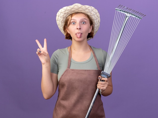 Photo gratuite impressionné jeune jardinier femme slave portant chapeau de jardinage sort la langue tenant le râteau à feuilles et gestes signe de la victoire sur violet