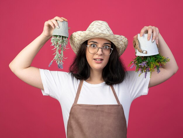 Impressionné jeune jardinière brune à lunettes optiques et en uniforme portant chapeau de jardinage détient des pots de fleurs à l'envers isolé sur un mur rose avec espace copie