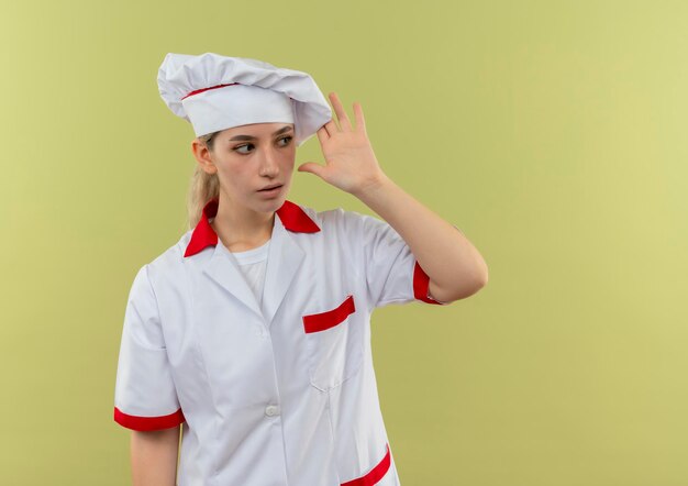 Impressionné jeune joli cuisinier en uniforme de chef regardant le côté mettant la main près de la tête regardant le côté isolé sur le mur vert avec espace de copie