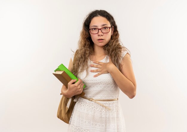 Impressionné jeune jolie écolière portant des lunettes et sac à dos tenant des livres mettant la main sur la poitrine isolé sur blanc