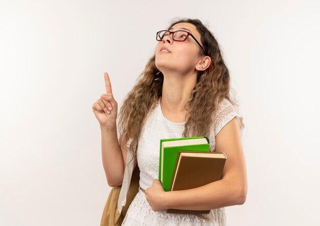 Impressionné jeune jolie écolière portant des lunettes et sac à dos tenant des livres à la recherche et pointant vers le haut isolé sur blanc