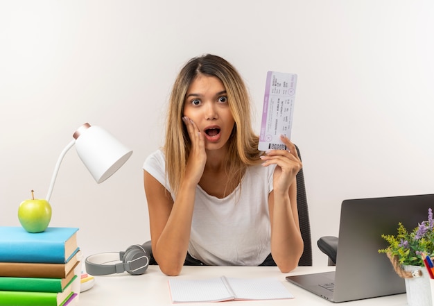 Impressionné jeune jolie étudiante assise au bureau avec des outils scolaires mettant la main sur la joue et tenant un billet d'avion isolé sur blanc