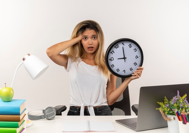 Impressionné jeune jolie étudiante debout derrière le bureau avec des outils scolaires mettant la main sur la tête et tenant horloge isolé sur blanc