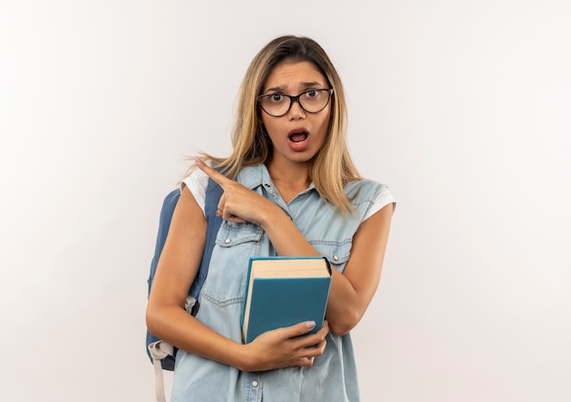 Impressionné jeune jolie étudiante portant des lunettes et sac à dos tenant le livre pointant sur le côté isolé sur blanc
