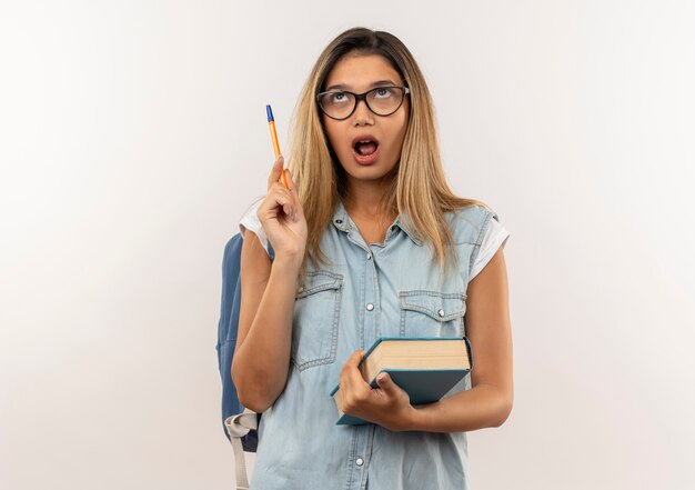 Impressionné jeune jolie étudiante portant des lunettes et sac à dos tenant un livre et un stylo en levant isolé sur blanc