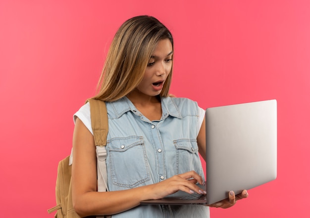 Photo gratuite impressionné jeune jolie étudiante portant un sac à dos à l'aide et en regardant un ordinateur portable isolé sur rose