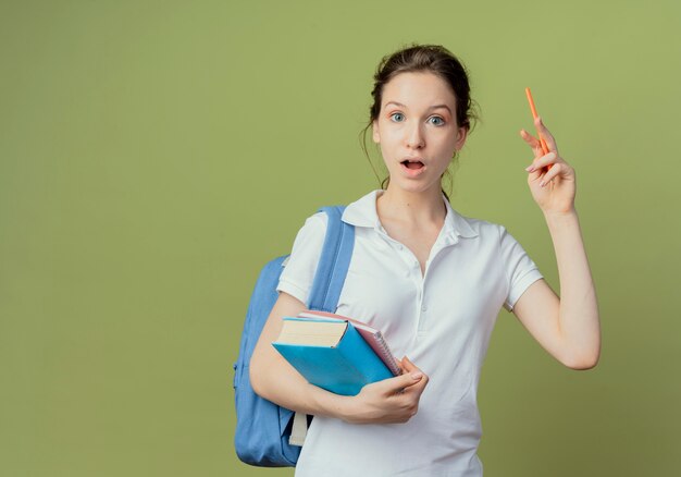 Impressionné jeune jolie étudiante portant sac à dos tenant bloc-notes et livre soulevant un stylo isolé sur fond vert olive avec espace de copie