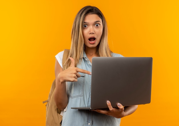 Impressionné jeune jolie étudiante portant sac à dos tenant et pointant sur ordinateur portable isolé sur orange