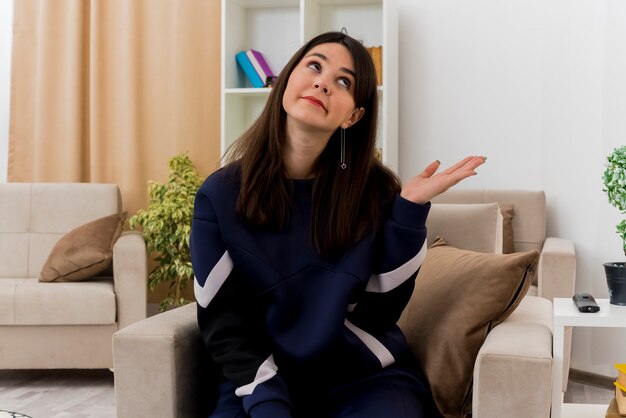 Impressionné jeune jolie femme caucasienne assise sur un fauteuil dans un salon conçu à la recherche de côté montrant la main vide