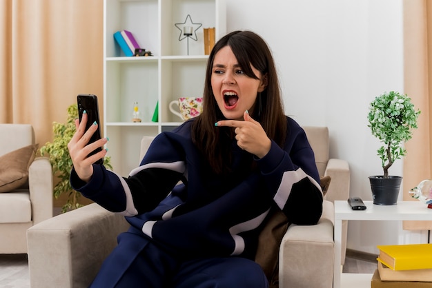 Impressionné jeune jolie femme caucasienne assise sur un fauteuil dans le salon conçu tenant et regardant le téléphone mobile et pointant vers elle