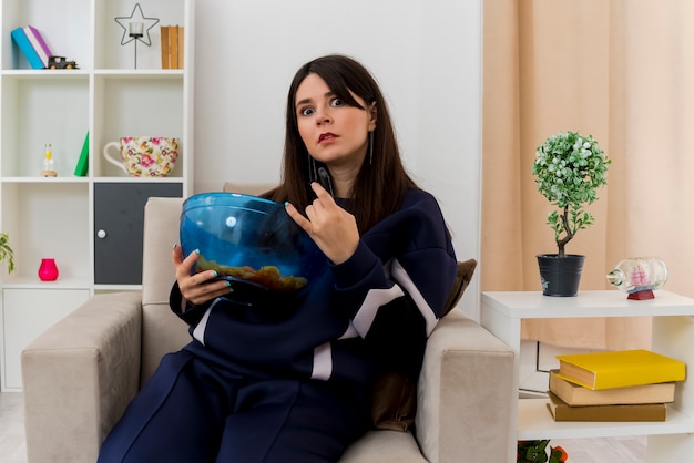 Impressionné jeune jolie femme caucasienne assise sur un fauteuil dans un salon conçu tenant la télécommande et un bol de chips et faisant le geste de la roche