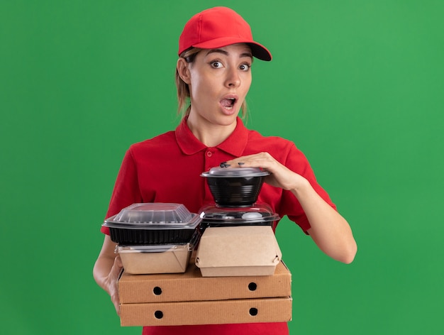 Impressionné jeune jolie femme de livraison en uniforme détient des emballages alimentaires en papier et des conteneurs sur des boîtes de pizza isolé sur mur vert