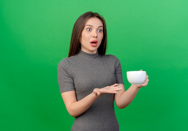 Impressionné jeune jolie femme tenant la tasse à la droite et montrant la main vide isolée sur fond vert avec espace copie