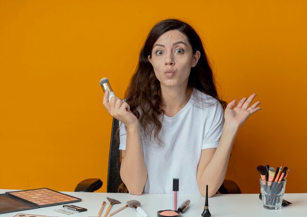 Impressionné jeune jolie fille assise à la table de maquillage avec des outils de maquillage en gardant la main dans l'air et tenant le pinceau de fondation avec de la crème de fondation mis sur son visage isolé sur fond orange