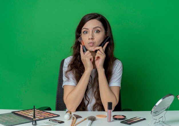 Impressionné jeune jolie fille assise à la table de maquillage avec des outils de maquillage tenant des pinceaux blush et poudre et toucher le visage avec eux isolé sur fond vert