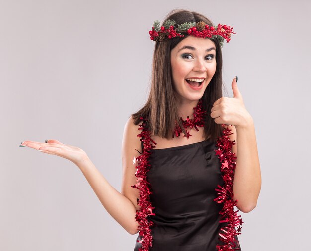 Impressionné jeune jolie fille caucasienne portant une couronne de Noël et une guirlande de guirlandes autour du cou montrant la main vide et le pouce vers le haut en regardant la caméra isolée sur fond blanc